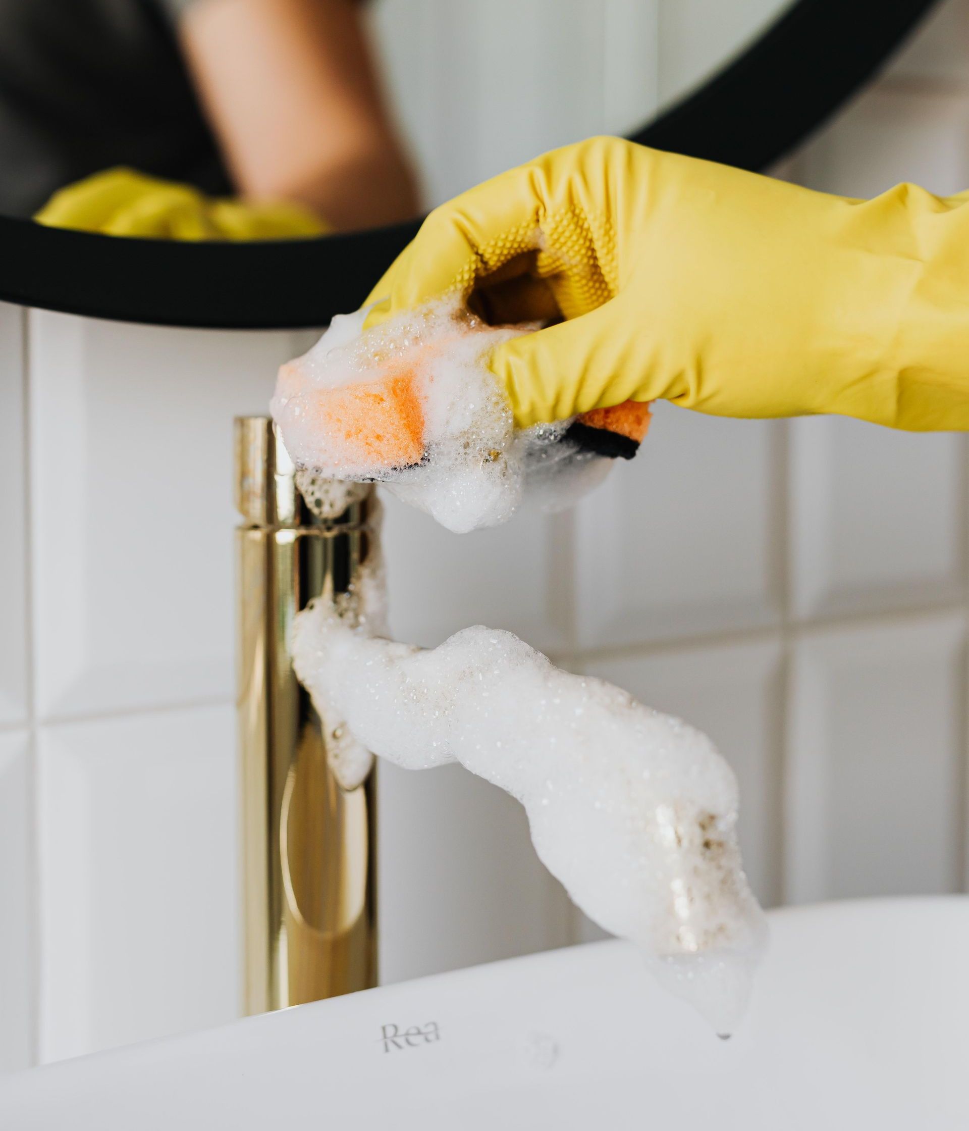 Anonymous person washing golden shiny faucet and sink in bathroom while wearing yellow latex glove during household care routine work